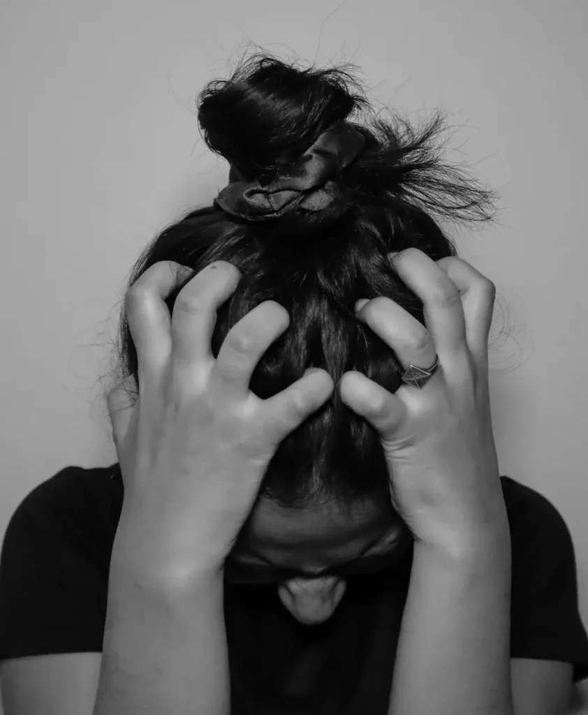 A close-up black and white photograph of a woman holding her head in emotional overwhelm, illustrating the hidden patterns behind emotional triggers behavior.