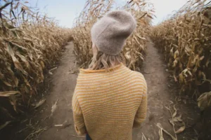 A woman seen from behind standing at a literal crossroads inside a tall corn maze, symbolizing the mental process of how to change your identity when you feel stuck.