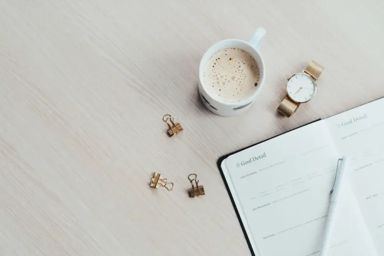 A minimalist overhead workspace view of an open goal-setting planner, a white cup of coffee, and a gold watch on a clean light wood desk.