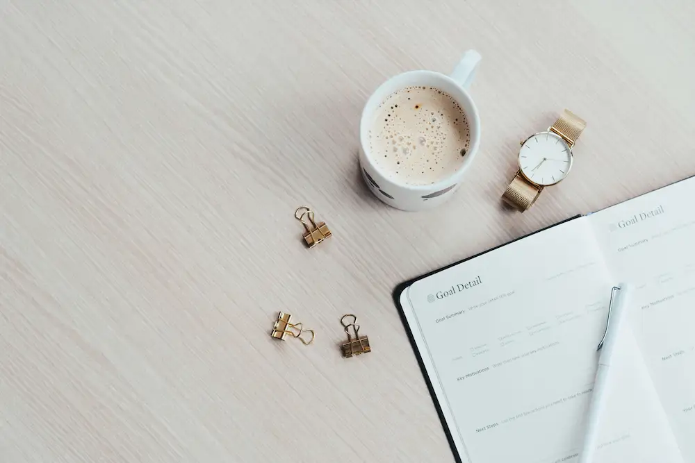 A minimalist overhead workspace view of an open goal-setting planner, a white cup of coffee, and a gold watch on a clean light wood desk.