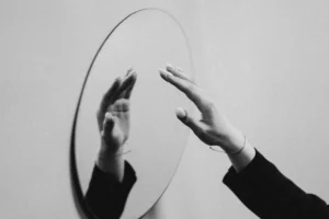 Hand reaching toward its reflection in a round mirror, symbolizing self-talk, affirmations, and subconscious belief change.