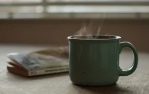 A green ceramic mug with steam visibly rising from a hot drink, sitting on a wooden desk. An out-of-focus paperback book lies flat to its left in a softly lit room.