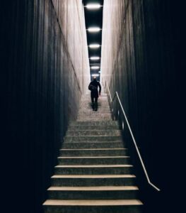 A person climbing a tall, narrow set of concrete stairs in a dimly lit, minimalist modern building, symbolizing progressive mental conditioning.