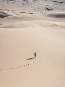 Person walking across sand dunes along a trail of footprints, representing old habits and repeated behavior patterns