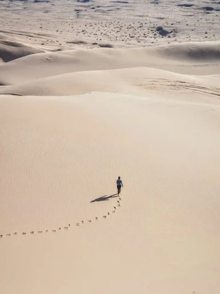 Person walking across sand dunes along a trail of footprints, representing old habits and repeated behavior patterns