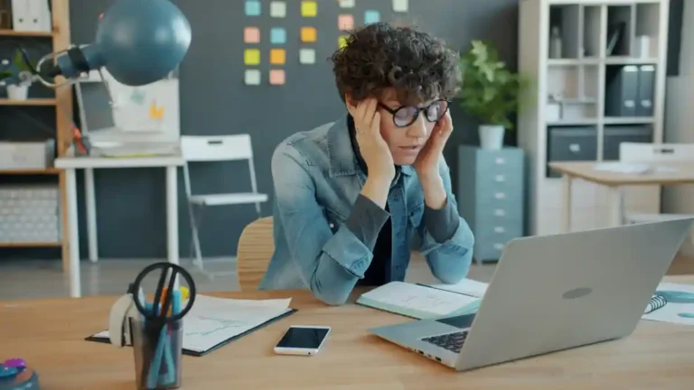 Woman sitting at her desk with her hands against her head, appearing frustrated while looking at a laptop.