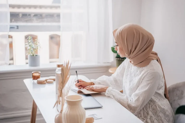 Woman writing in a notebook at a sunlit desk beside a window.